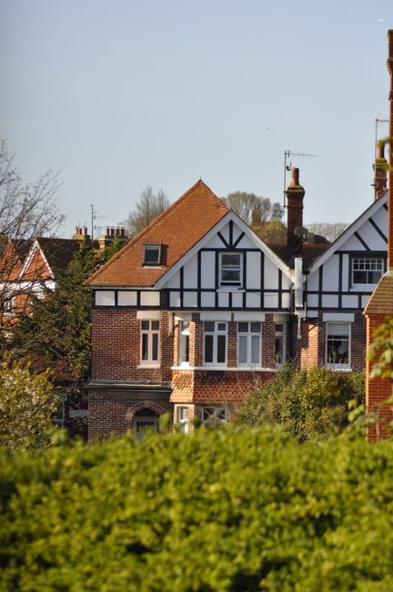 A row of traditional British houses with brick and timber-framed facades, featuring pitched roofs with terracotta tiles, white window frames, and chimneys, set against a clear blue sky. The foreground