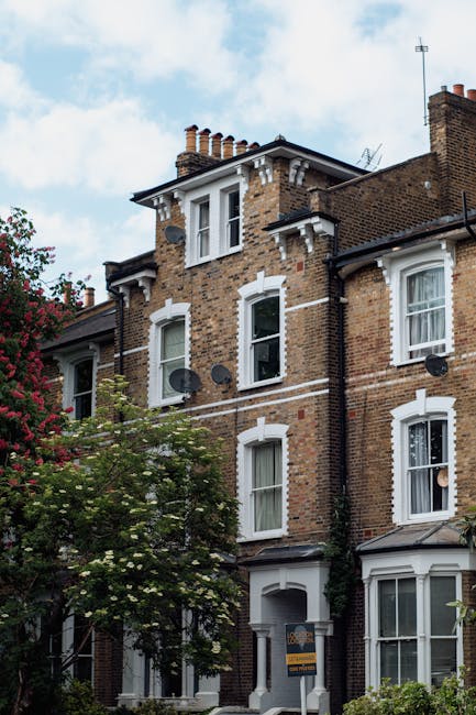 A row of traditional British houses with brick and timber-framed facades, featuring pitched roofs with terracotta tiles, white window frames, and chimneys, set against a clear blue sky. The foreground