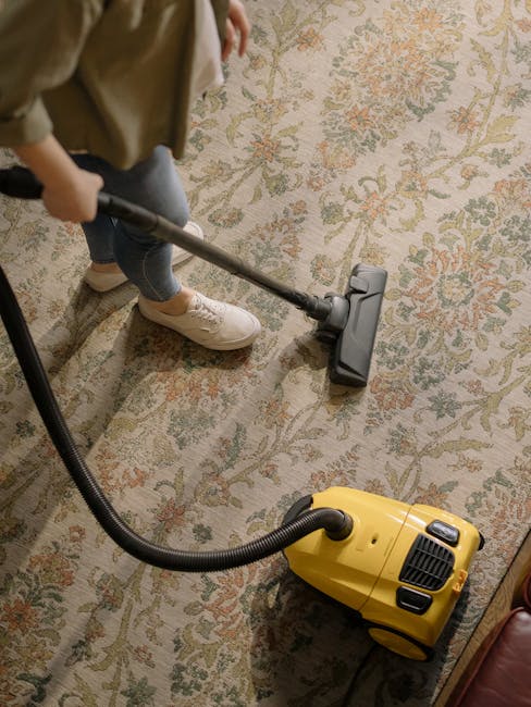 A young boy using a portable vacuum cleaner to perform surface cleaning on a beige fabric sofa in a bright living room, with an older man lying relaxed on the sofa, wearing casual grey and orange clot