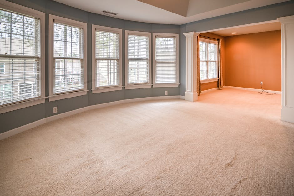 A spacious living room with a light beige carpet that appears clean and well-maintained. The room features multiple large windows framed with white trim, allowing natural light to brighten the space.