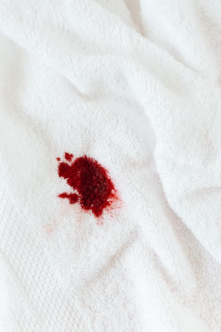 A close-up view of a white cloth garment, likely a shirt or dress, with a noticeable red stain resembling blood splatter, placed against a soft pink background. The fabric appears clean except for the