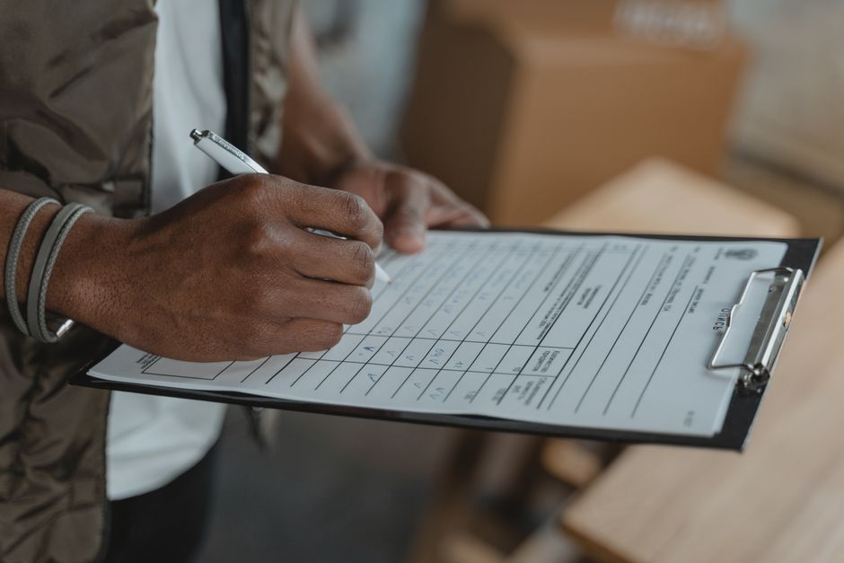 Close-up of a person's hand holding a silver pen and filling out a cleaning inspection or maintenance checklist on a black clipboard, with visible printed rows and columns. The individual is wearing a